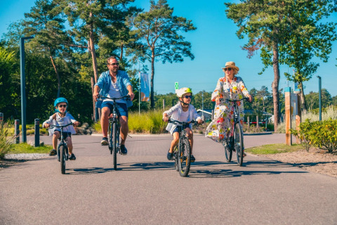 En familie cykler sammen på en solrig dag i en feriepark med glamping-indkvartering og grønne omgivelser.