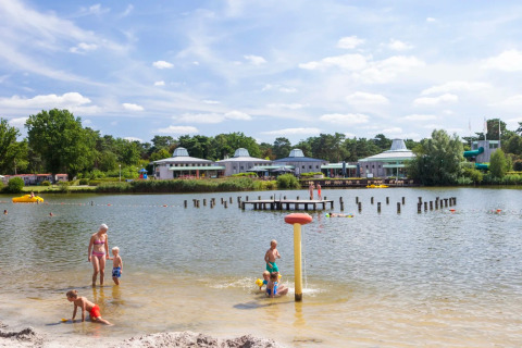 Familien genießen einen Sommertag am Badesee eines Ferienparks mit Glamping, Kinder spielen im Wasser.