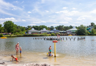 Familier nyder sommerdag ved søbred i feriepark med glampingmuligheder, børn leger i vand og sand.