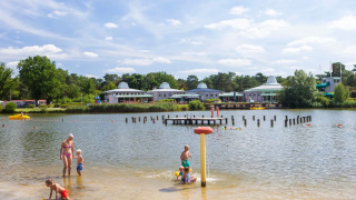 Familien genießen einen Sommertag am Badesee eines Ferienparks mit Glamping, Kinder spielen im Wasser.