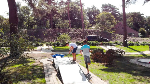 Two boys play mini-golf in a holiday park with lush greenery and glamping accommodations in the background.