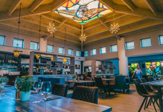 Elegant restaurant på et glamping resort med mosaikvinduer og farverigt glastag, varme træmøbler og planter.