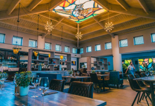 Elegant restaurant på et glamping resort med mosaikvinduer og farverigt glastag, varme træmøbler og planter.