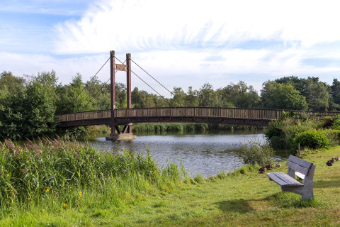 Pont en bois au-dessus d’un lac dans un parc de vacances avec glamping, entouré de verdure et d’un banc.