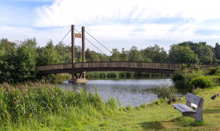 Holzbrücke über einen See im Ferienpark mit Glamping, umgeben von viel Grün und einer Bank am Ufer.
