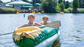 Zwei Kinder paddeln in einem grünen Schlauchboot auf einem See im Ferienpark mit Glamping-Angebot.