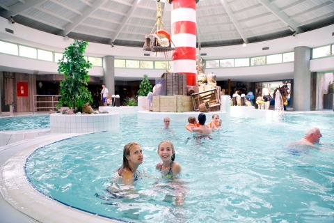 Indendørs swimmingpool i en feriepark med glamping, børn og voksne leger i vandet nær et fyrtårn.