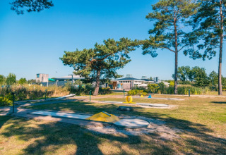 Udendørs minigolfbane ved et feriepark med glamping, omgivet af træer og moderne faciliteter i baggrunden.