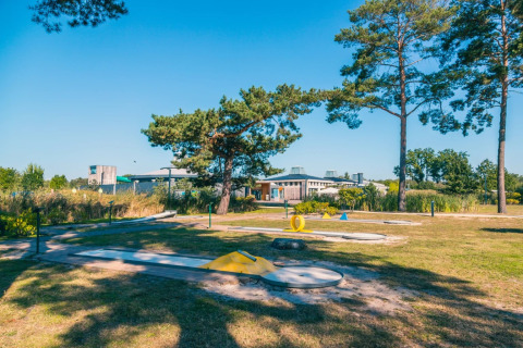 Buiten minigolfbaan bij een vakantiepark met glamping, omgeven door bomen en moderne gebouwen op de achtergrond.