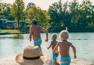 Famille courant vers le lac dans un parc de vacances avec glamping, entourée d’arbres par temps ensoleillé.