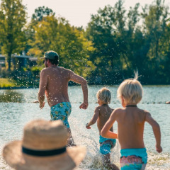 Familie läuft lachend ins Wasser an einem See im Ferienpark mit Glamping, umgeben von Bäumen.