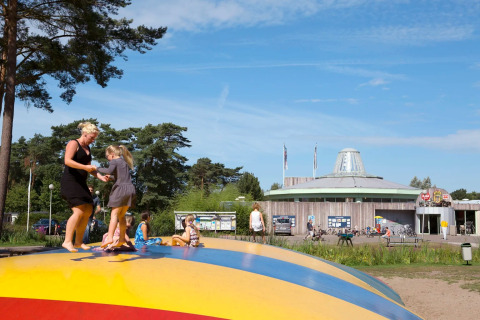 Adults and children play on an inflatable bouncer at a holiday park with a modern building and trees.