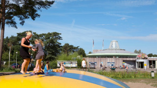 Kinder und Erwachsene spielen auf einem großen Hüpfkissen vor einem Ferienpark mit Bäumen und Fahrrädern.