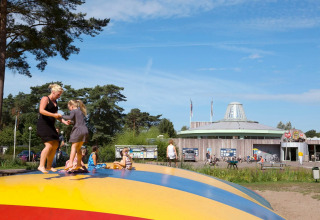 Adults and children play on an inflatable bouncer at a holiday park with a modern building and trees.
