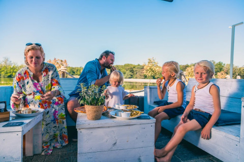 Familie isst zusammen im Freien auf weißen Holzbänken in einem Ferienpark mit glamping Unterkunft.