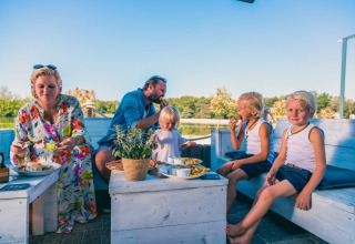 En familie nyder udendørs måltid ved et feriepark, omgivet af grøn natur, på lyse træbænke.