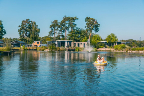 Two people paddle an inflatable boat on a lake with modern glamping accommodations at a holiday park.