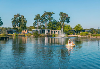 Two people paddle an inflatable boat on a lake with modern glamping accommodations at a holiday park.