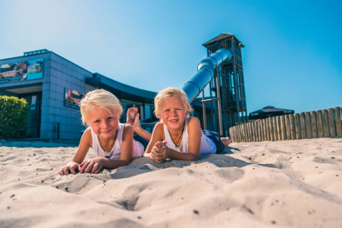 Zwei Kinder spielen im Sand vor einem modernen Gebäude und einer Rutsche in einem Glamping-Ferienpark.