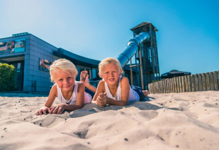 To børn leger i sandet foran en moderne bygning og vandrutsjebane i en feriepark med glampingmuligheder.