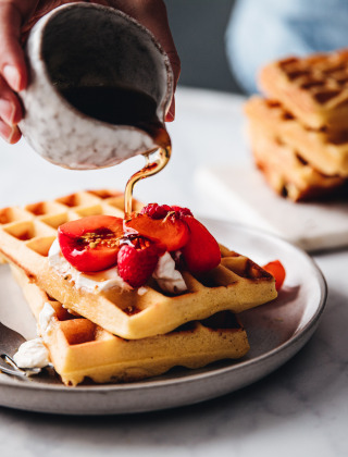 Una mano vierte sirope sobre gofres con frutas y crema en un plato de desayuno, listo para comer.