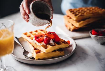 Sirup wird über frisch gebackene Waffeln mit Beeren und Sahne auf einem Frühstücksteller gegossen.