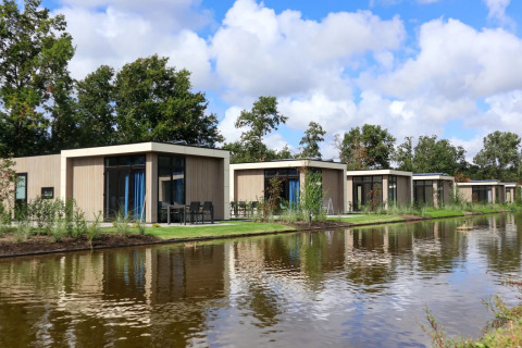 Row of modern glamping lodges by a lake in a holiday park, surrounded by trees and blue sky.
