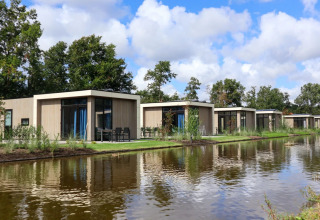 Rij moderne glampingchalets langs het water in een vakantiepark met veel groen en blauwe lucht.