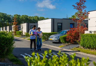 A young couple walk past modern glamping cabins surrounded by greenery in a holiday park on a sunny day.