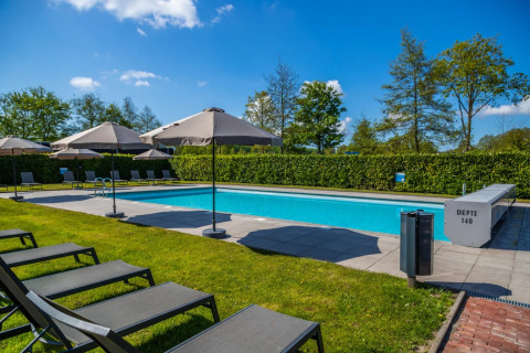 Buitenzwembad op een vakantiepark met ligstoelen, parasols en groene hagen onder een strakblauwe lucht.