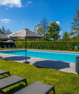 Piscina exterior en un parque vacacional con tumbonas, sombrillas y setos bajo un cielo azul despejado.