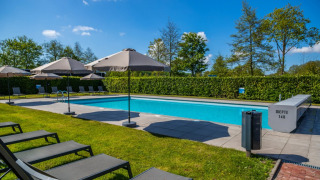 Piscina exterior en un parque vacacional con tumbonas, sombrillas y setos bajo un cielo azul despejado.