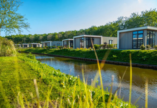 Moderne glampinglodges in een vakantiepark aan een kanaal, met veel groen, bomen en een heldere hemel.
