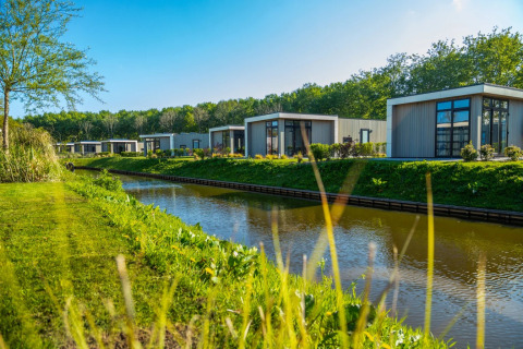 Moderne glampinglodges in een vakantiepark aan een kanaal, met veel groen, bomen en een heldere hemel.