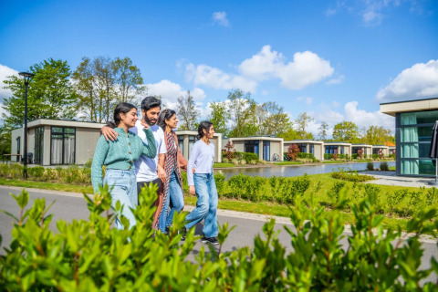 A group of people walking happily past modern glamping lodges by a pond at a holiday park resort.