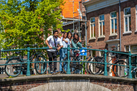 En gruppe unge mennesker nyder en solrig dag på en bro med cykler i en hollandsk by med klassisk arkitektur.