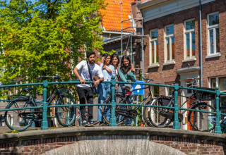 A group of young people enjoy a sunny day on a bridge with bicycles in a Dutch city with classic houses.
