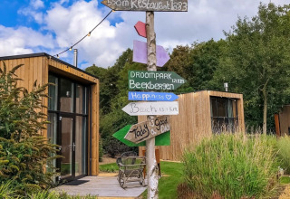 Foto bij een vakantiepark met moderne glampinghutten, kleurrijke wegwijzers en groene omgeving in de zon.