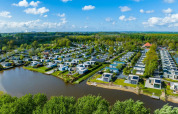 Luftfoto af en feriepark med moderne glampingboliger ved søbred og omgivet af grøn natur og skov.