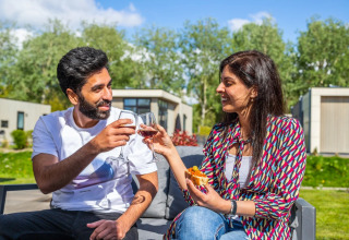 Koppel geniet van wijn en hapjes buiten bij een glamping vakantiepark met veel groen en chalets.