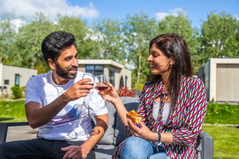 Paar genießt Wein und Snacks auf der Terrasse in einem Glamping-Ferienpark mit grüner Umgebung.