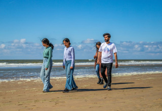 Vier mensen wandelen op het strand bij een vakantiepark met glamping, tijdens een zonnige dag aan zee.
