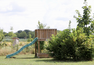 Een speeltuin met groene glijbaan en houten structuur tussen struiken bij Camping de Heerlijkheid Vorenseinde.