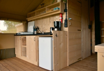 Interieur van een safaritent met compacte houten keuken, gasfornuis, koffiezetapparaat en minikoelkast.