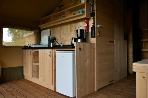 Binnenzicht in een safaritent met een houten keukenblok, gasfornuis, koffiezet en kleine koelkast.