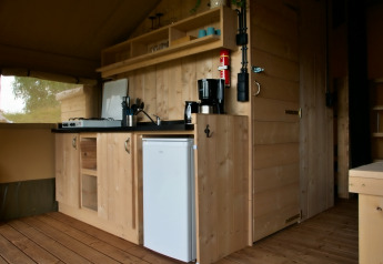 Interieur van een safaritent met compacte houten keuken, gasfornuis, koffiezetapparaat en minikoelkast.
