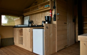 Interior view of a safari tent featuring a compact wood kitchen, gas stove, coffee maker, and mini fridge.