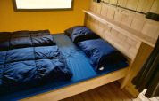 Gezellige glamping accommodatie met houten bedframe, donkerblauw beddengoed en raam op de achtergrond.