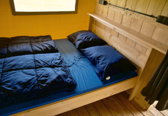 Gezellige glamping accommodatie met houten bedframe, donkerblauw beddengoed en raam op de achtergrond.