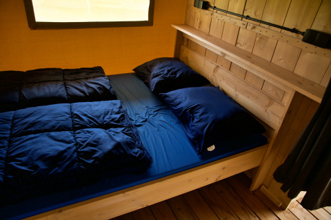 Gezellige glamping accommodatie met houten bedframe, donkerblauw beddengoed en raam op de achtergrond.